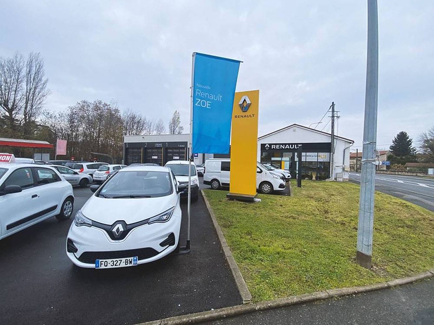 Devant une concession Renault, une Renault Zoe blanche est garée près d'un drapeau bleu Nouvelle Renault ZOE et d'un totem jaune Renault.