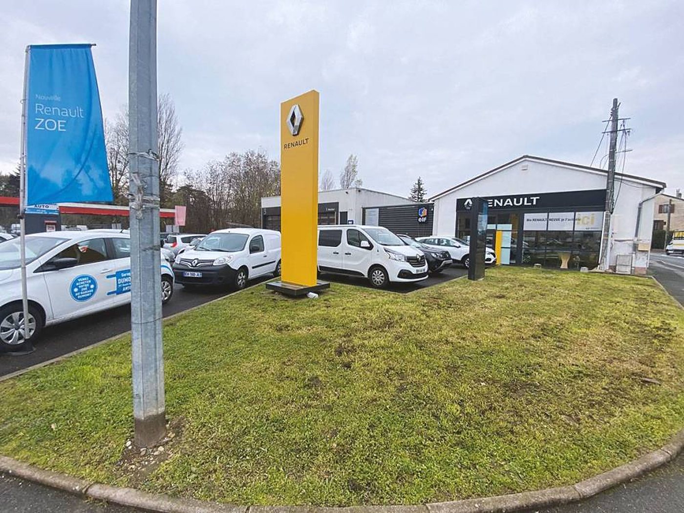 Façade d'une concession Renault avec son totem jaune, une banderole bleue Renault ZOE et plusieurs véhicules blancs garés sur le parking.