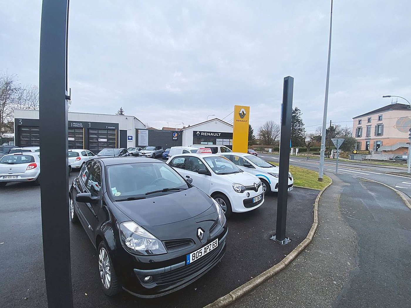 Devant une concession Renault, des voitures noires et blanches sont garées sur le parking près de l'atelier et du totem jaune.