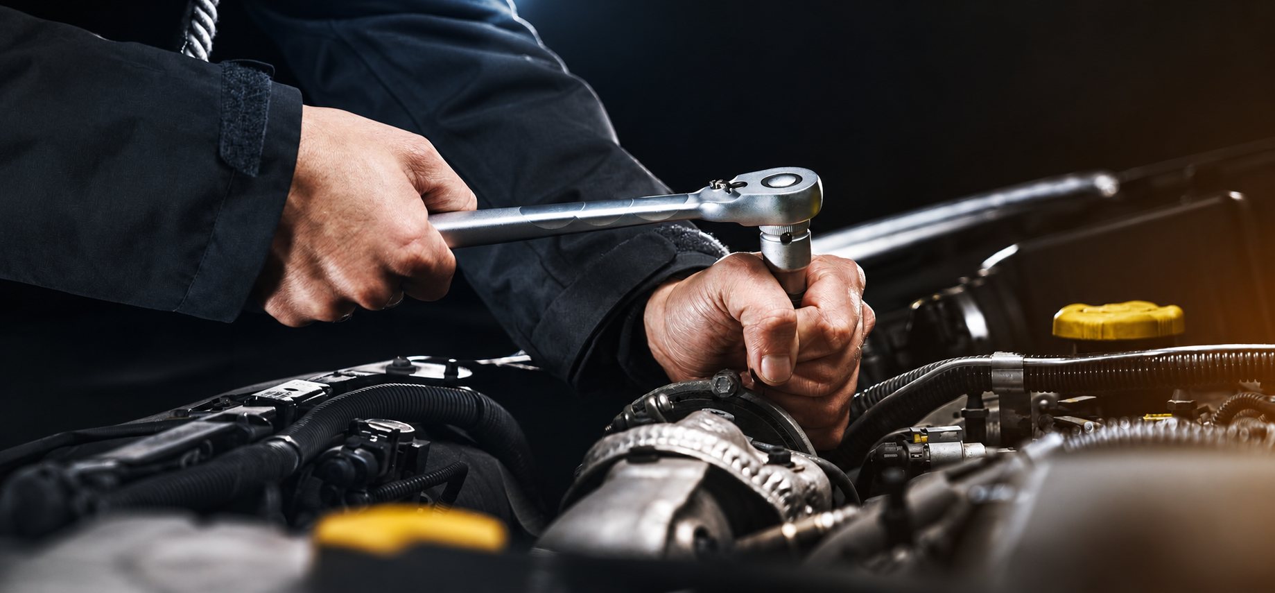 Les mains d'un mécanicien utilisent une clé à cliquet pour intervenir sur le moteur d'une voiture au capot ouvert.