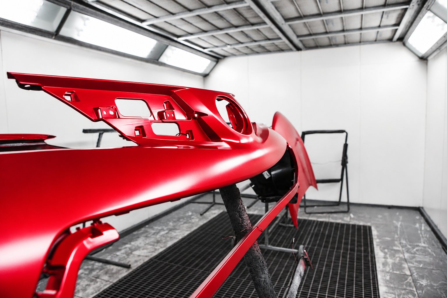 Un pare-chocs de voiture peint en rouge mat est suspendu sur un support à l'intérieur d'une cabine de peinture blanche au sol grillagé.
