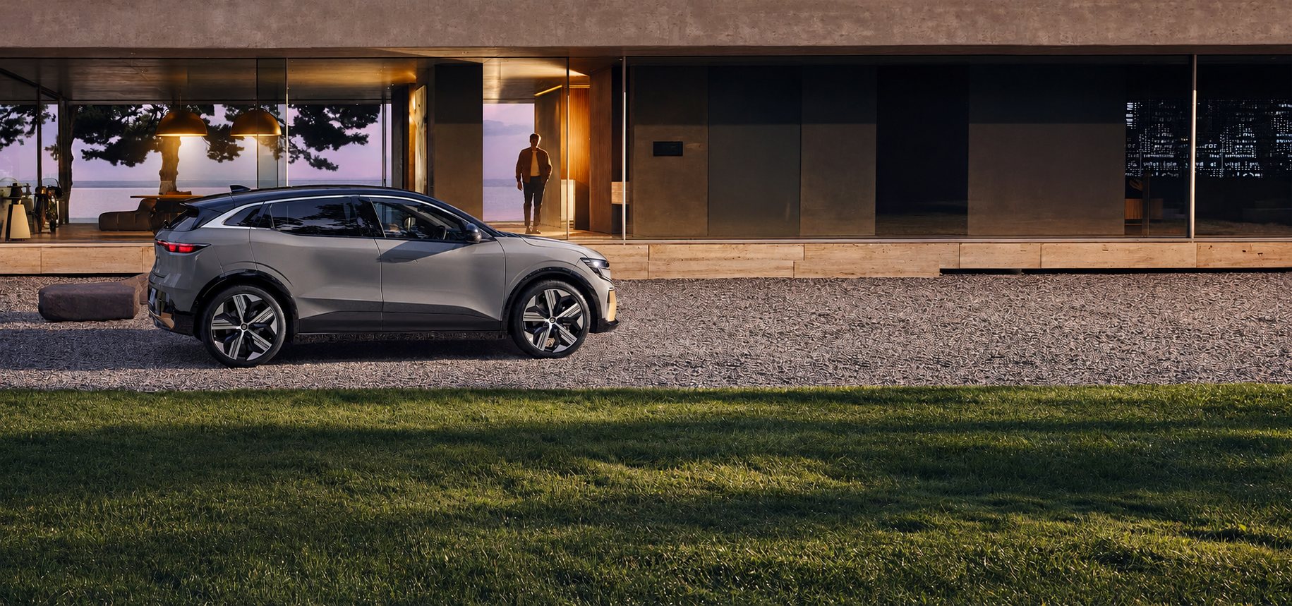 Un SUV électrique gris est garé sur du gravier devant une maison moderne à grandes baies vitrées, tandis qu'un homme se tient à l'intérieur au crépuscule.