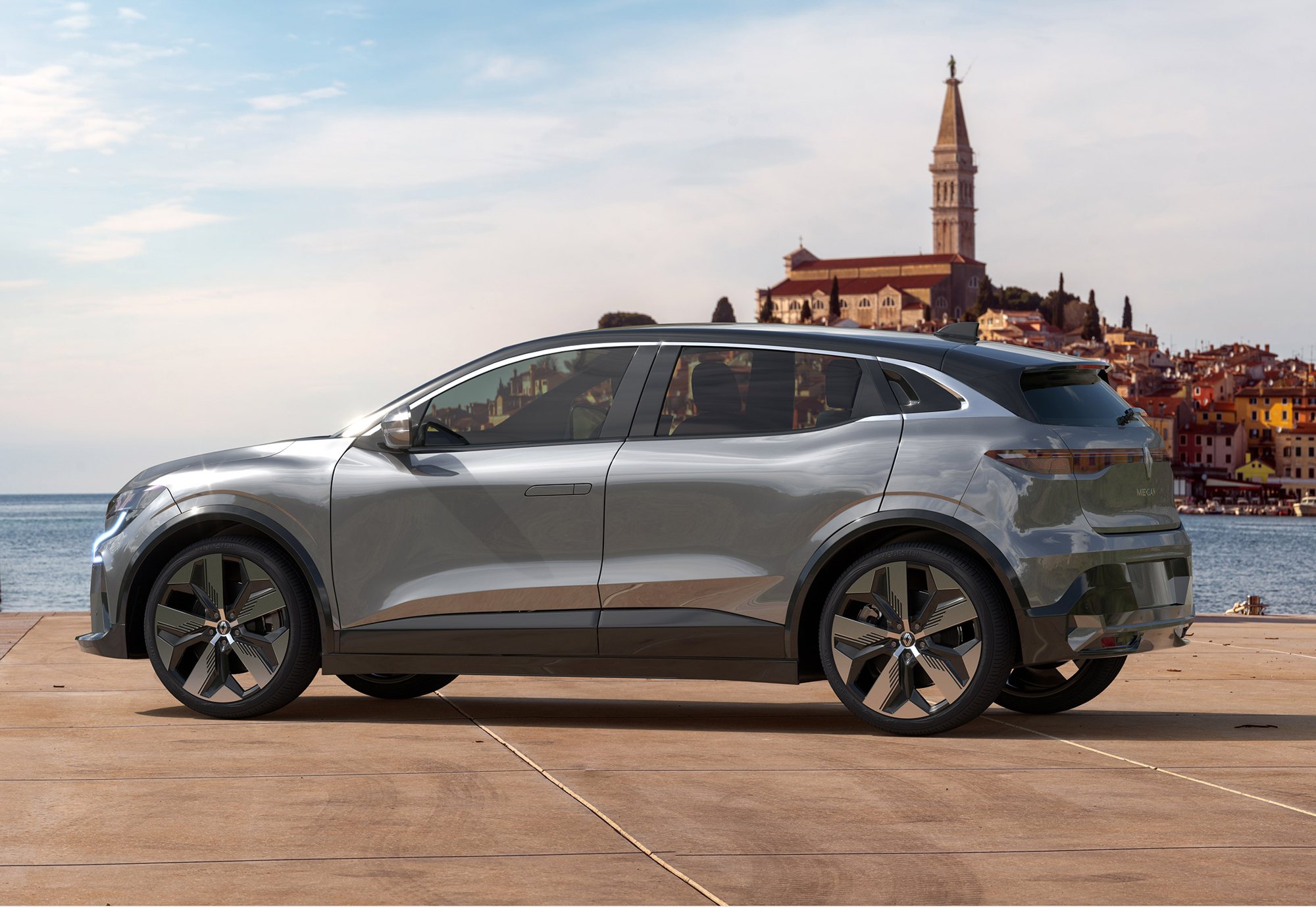 Un SUV électrique gris métallisé est garé sur une esplanade face à la mer, avec une ville côtière colorée et son clocher en arrière-plan.